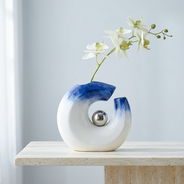 A modern ceramic vase with a white and blue gradient design, featuring a central metallic silver sphere and a split opening holding a white orchid stem, displayed on a light stone table against a soft blue-gray background.