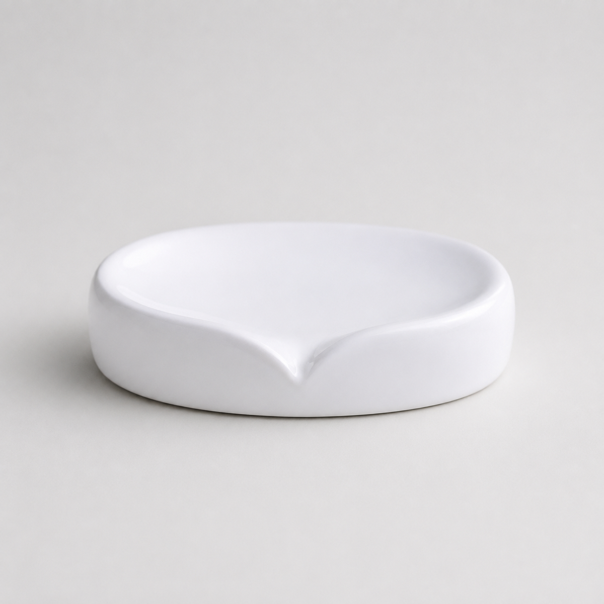 A minimalist white ceramic soap dish with a small pouring spout, photographed on a clean, light gray background.
