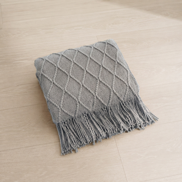 A square gray knit throw pillow with a diamond lattice pattern and fringed edges, placed on a light beige wood floor.