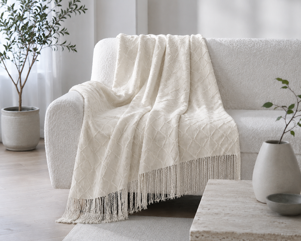 An beige textured throw blanket with fringe trim draped over a white boucle sofa in a bright, modern living room setting.