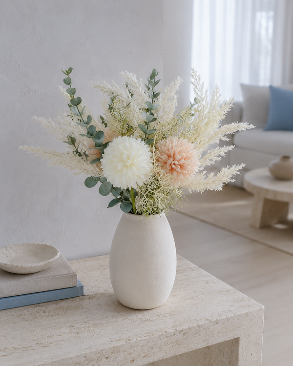 A dried floral arrangement in a matte white ceramic vase, placed on a light stone side table in a bright, modern living room.