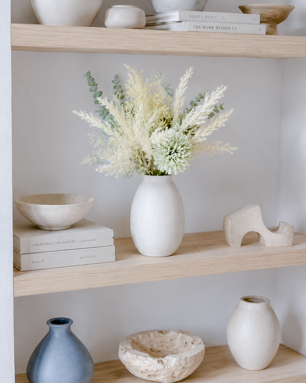 A styled Scandinavian interior shelf display with a white ceramic vase holding a dried floral arrangement, surrounded by matte ceramic bowls, a small blue vase, a stone bowl, and books on light wood shelves.