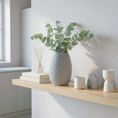 A textured light gray ceramic vase filled with eucalyptus branches, styled on a light wood floating shelf next to books, a reed diffuser, and small ceramic containers in a bright, modern room.