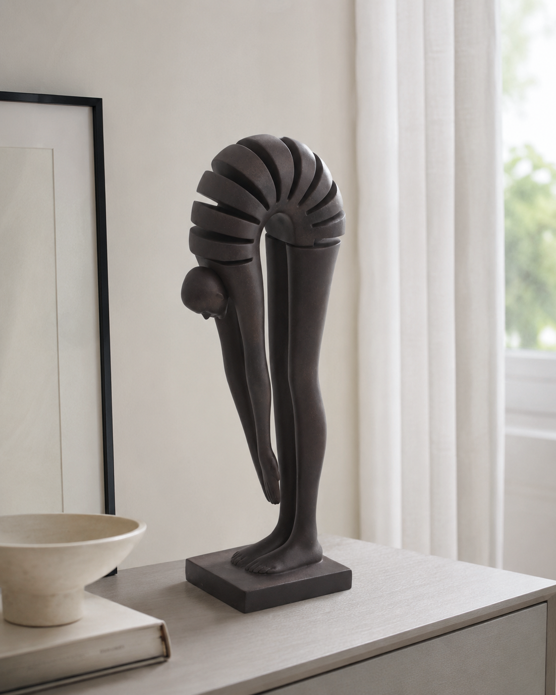 A dark charcoal gray abstract sculpture of a stylized human figure in a bent-over pose, displayed on a light gray console table in a bright, modern room next to a window with white curtains.