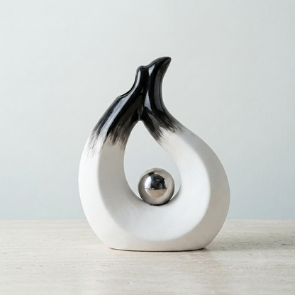 A modern abstract ceramic sculpture vase of a stylized flame in black and white, with a polished silver metallic sphere nestled in its curved body, set against a neutral light gray background.