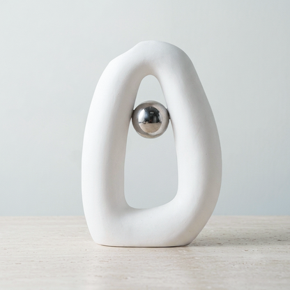 A white abstract ring-shaped sculpture with a polished silver metallic ball suspended in the center, displayed on a light wood surface against a soft light gray background.