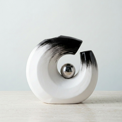 A modern abstract ceramic vase in a spiral C-shape, featuring a glossy black and white gradient finish with a polished metallic silver sphere at its center, displayed on a light-colored surface against a neutral background.