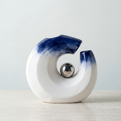 An abstract ceramic vaase in a C-shape with a white body and dark blue gradient glaze, featuring a polished metallic silver sphere inside the hollow center, set against a light gray background.