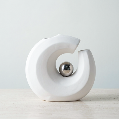 A modern white ceramic C-shaped ceramic vase with a polished silver metallic ball in the center, set on a light wooden surface against a neutral light gray background.
