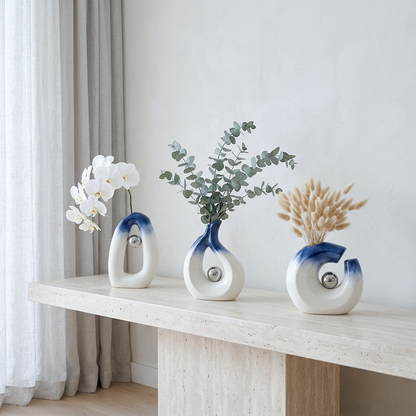 Three ceramic spiral-shaped vases with blue gradient accents and silver circular details, displayed on a light stone console table. The vases are styled with white orchids, eucalyptus branches, and dried pampas grass against a neutral wall and curtain background.