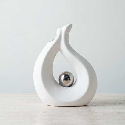 A white ceramic decorative sculpture with a smooth, flowing abstract shape and a polished silver metallic ball centered inside, set on a light stone surface against a soft light gray background.