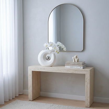 A light beige stone console table in a minimalist Scandinavian room, styled with a white sculptural vase, white orchids, a small white animal figurine, and a stack of books beneath an arched wall mirror.