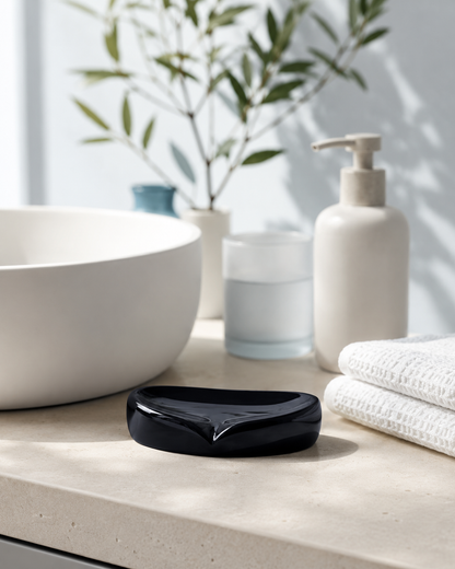 A glossy black soap dish with a curved, modern shape resting on a light beige stone countertop in a bathroom setting, with a white vessel sink, a cream-colored pump bottle, and folded white towels in the background.