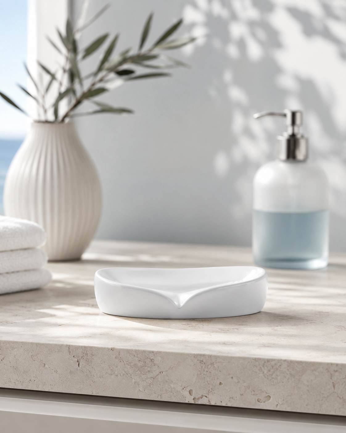 A white ceramic soap dish with a curved, leaf-like notch sits on a light stone bathroom counter. In the softly blurred background, a white ribbed vase with green stems, folded white towels, and a frosted glass pump bottle are visible.