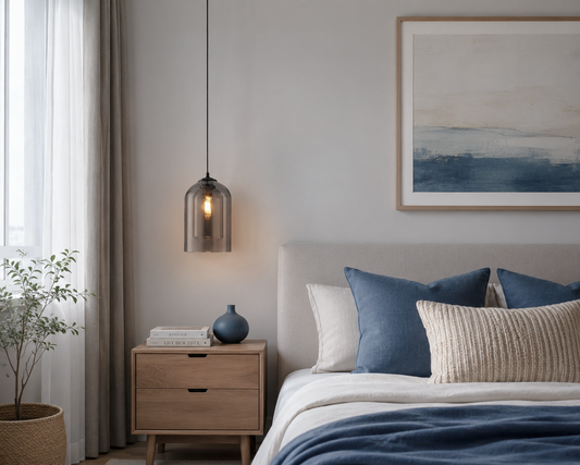 A modern bedroom scene featuring a dark gray glass pendant light hanging over a light wood nightstand next to a bed with blue and cream pillows.