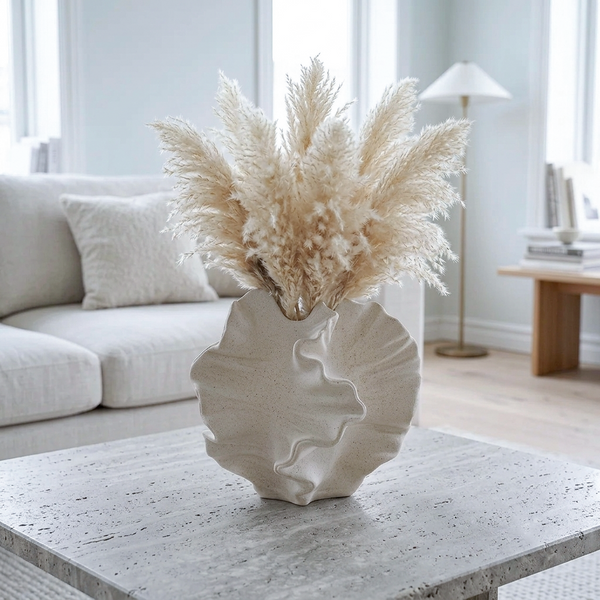 A sculptural, textured off-white ceramic vase filled with fluffy beige pampas grass, set on a speckled stone coffee table in a bright, modern living room with a light gray sofa and floor lamp in the background.