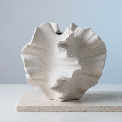A sculptural ceramic vase with a ruffled, shell-like silhouette in a warm off-white speckled finish, displayed on a matching stone slab against a soft light blue-gray background.