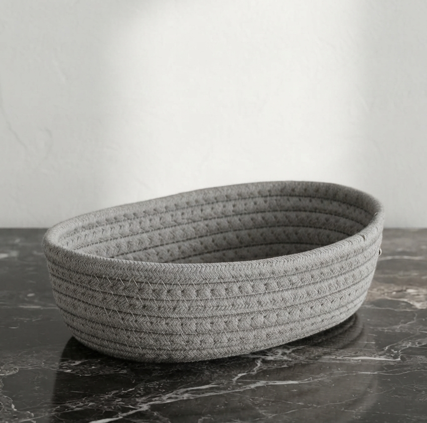 A gray woven rope basket with a textured basketweave pattern, resting on a dark marble surface against a light gray wall.