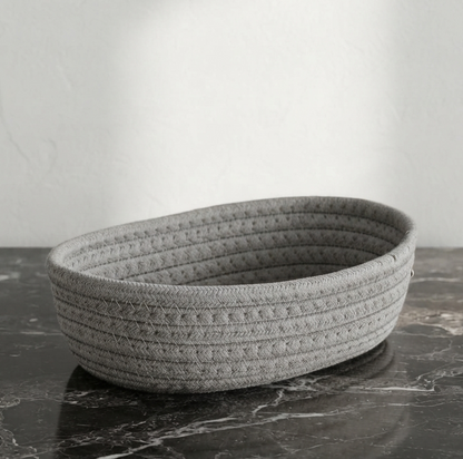 A gray woven rope basket with a textured basketweave pattern, resting on a dark marble surface against a light gray wall.