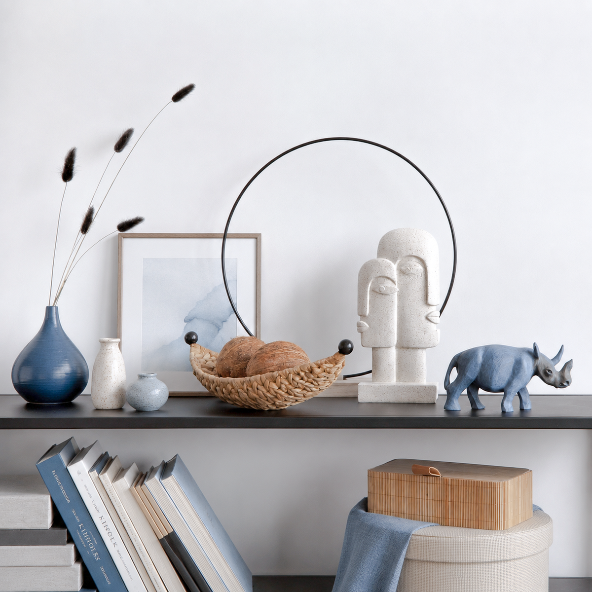 A styled interior shelf scene with decorative objects including a blue vase with dried stems, a framed print, a woven basket with bread, a white sculptural face lamp, a cerulean rhinoceros figurine, books, and a round box with a blue cloth.