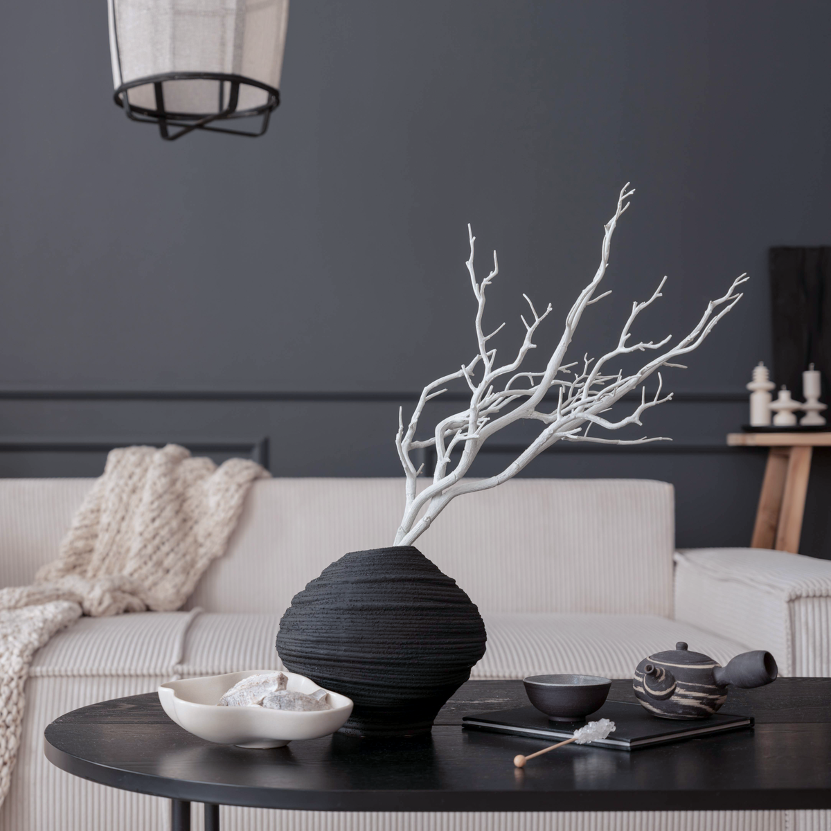A modern minimalist black ribbed vase holding a white decorative branch, styled on a dark round table in a contemporary living room setting.