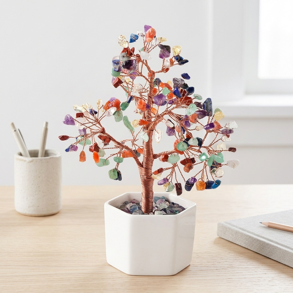 A copper wire crystal tree with colorful gemstone leaves in a white hexagonal pot, displayed on a light wood desk next to a cup with pens and a notebook.