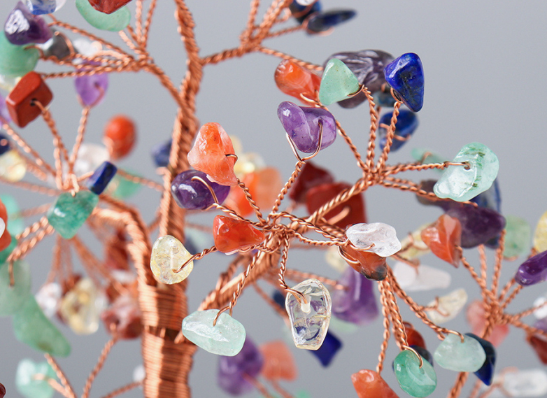 A close-up of a copper wire tree of life crystal jewelry tree with multiple colorful tumbled gemstones on a neutral gray background.
