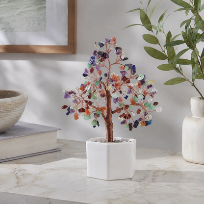 A crystal tree centerpiece with colorful gemstone leaves on a copper wire branch, set in a white square ceramic pot on a marble surface.