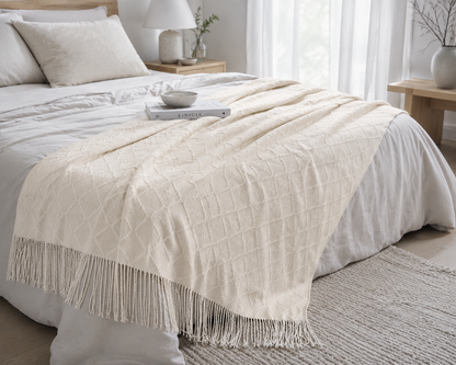 Abeige-colored textured throw blanket with fringe trim draped over a bed with white bedding in a bright, modern bedroom.