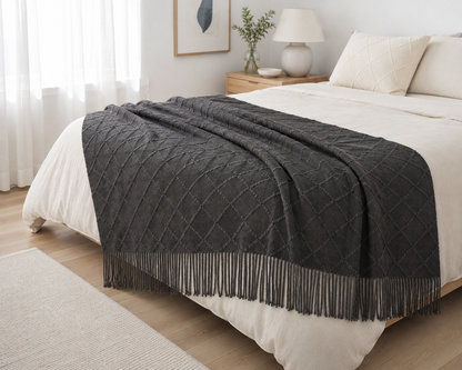 A dark charcoal gray textured throw blanket with fringe trim draped over a bed with white bedding in a bright, modern bedroom.