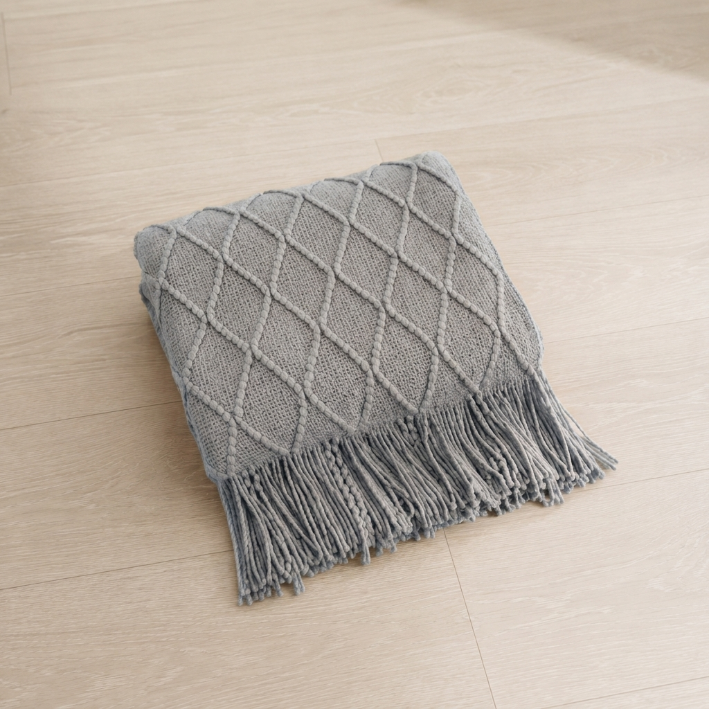 A square gray knit throw pillow with a diamond lattice pattern and fringed edges, placed on a light beige wood floor.