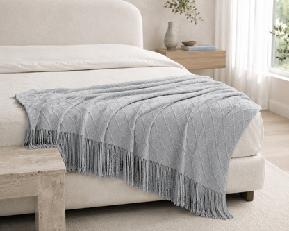 A light gray textured throw blanket with fringe trim draped over a cream-colored bed in a bright, modern bedroom setting.