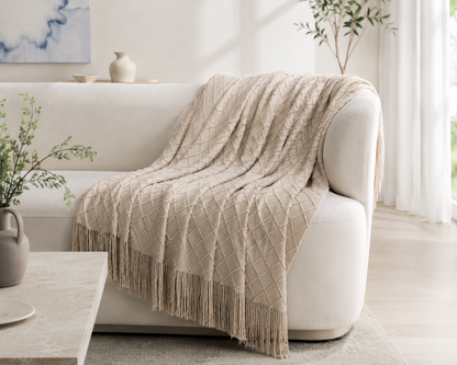 A beige textured knit throw blanket with fringe is draped over a cream-colored sofa in a bright, modern living room setting.