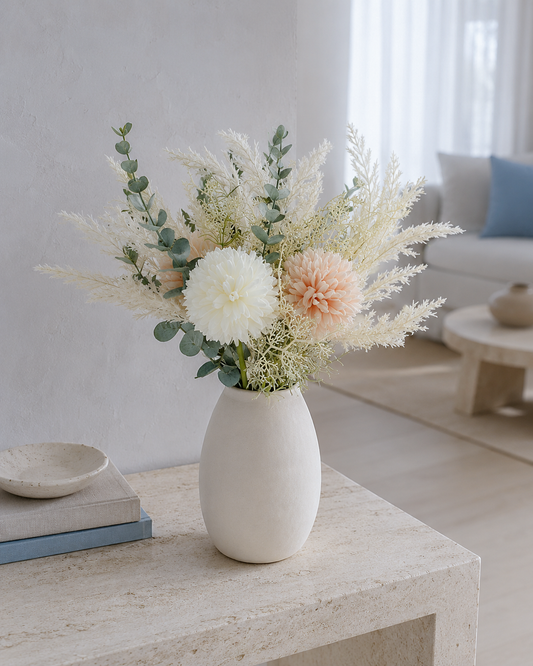 A dried floral arrangement in a matte white ceramic vase, placed on a light stone side table in a bright, modern living room.
