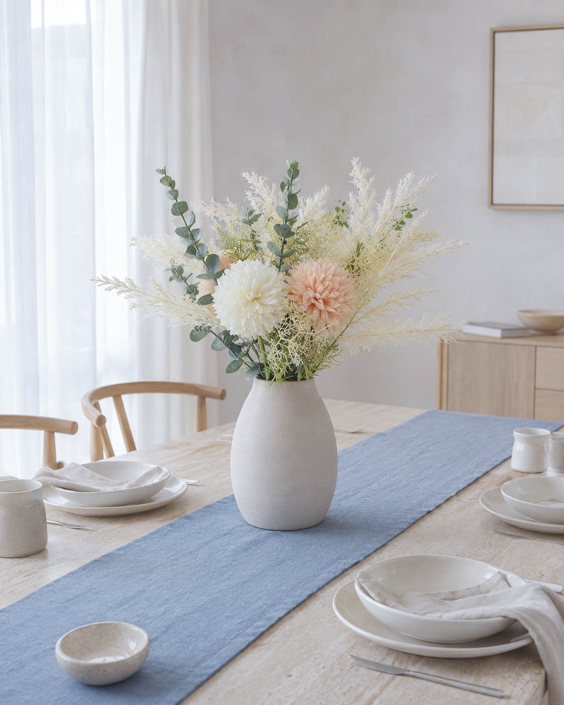 A white ceramic vase filled with a floral arrangement of white and blush pink flowers and green eucalyptus stems, set on a light wood dining table with a blue runner, white plates, and napkins in a softly lit room.