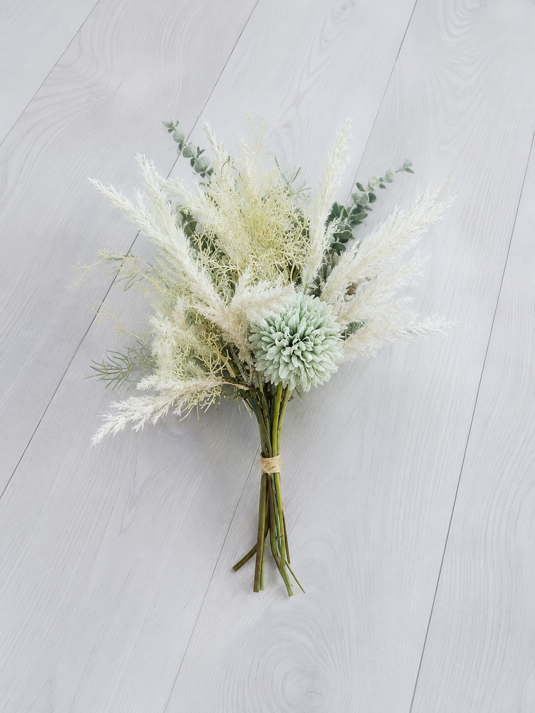 A dried floral arrangement featuring pampas grass, dusty millie, eucalyptus, and a pale green globe accent, tied with a natural twine band and placed on a light gray wood plank surface.