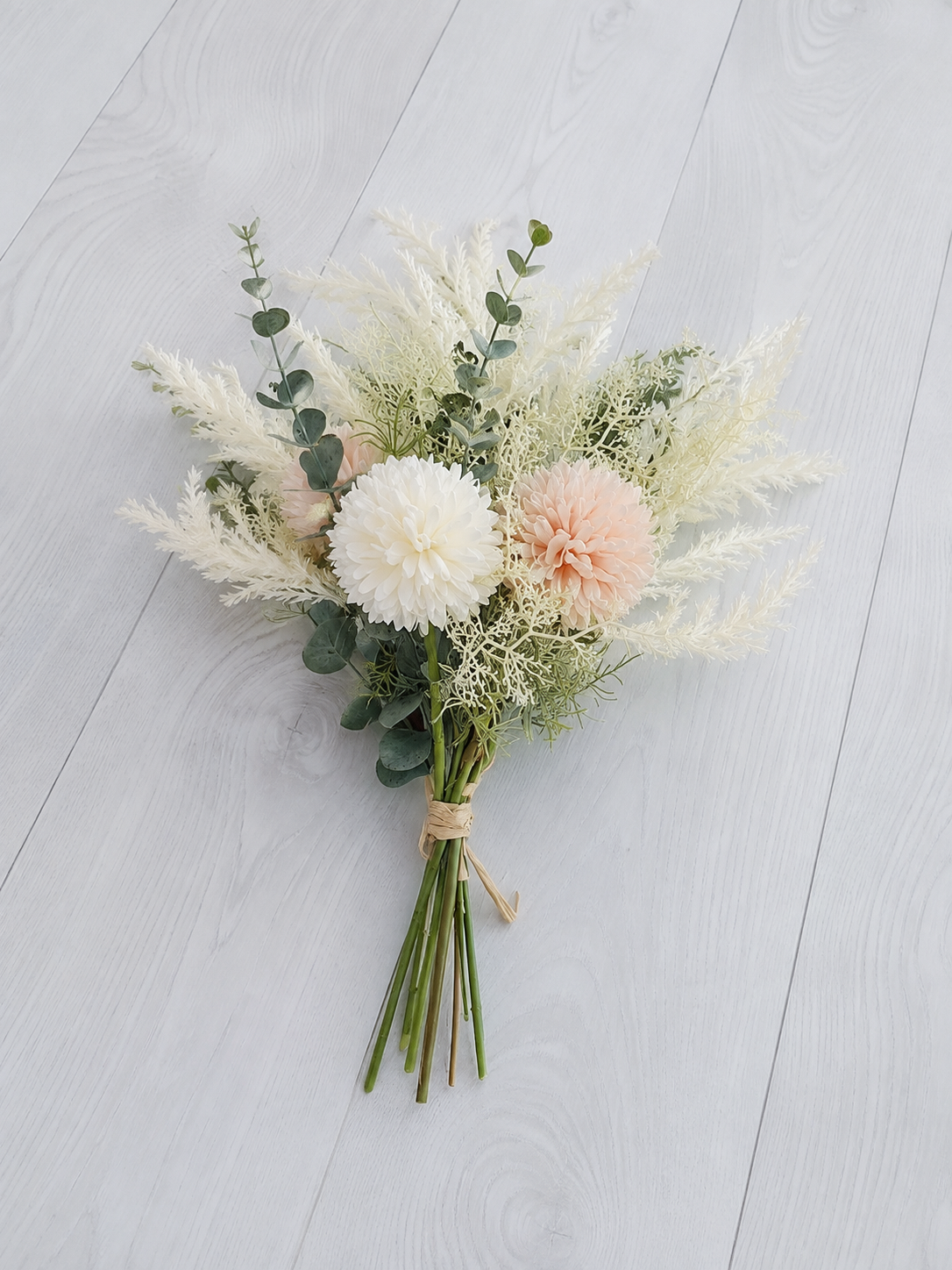 A dried floral arrangement featuring white and blush pink flowers, green eucalyptus stems, and fluffy white pampas grass, tied with a simple twine bow and photographed from above on a light gray wood plank background.