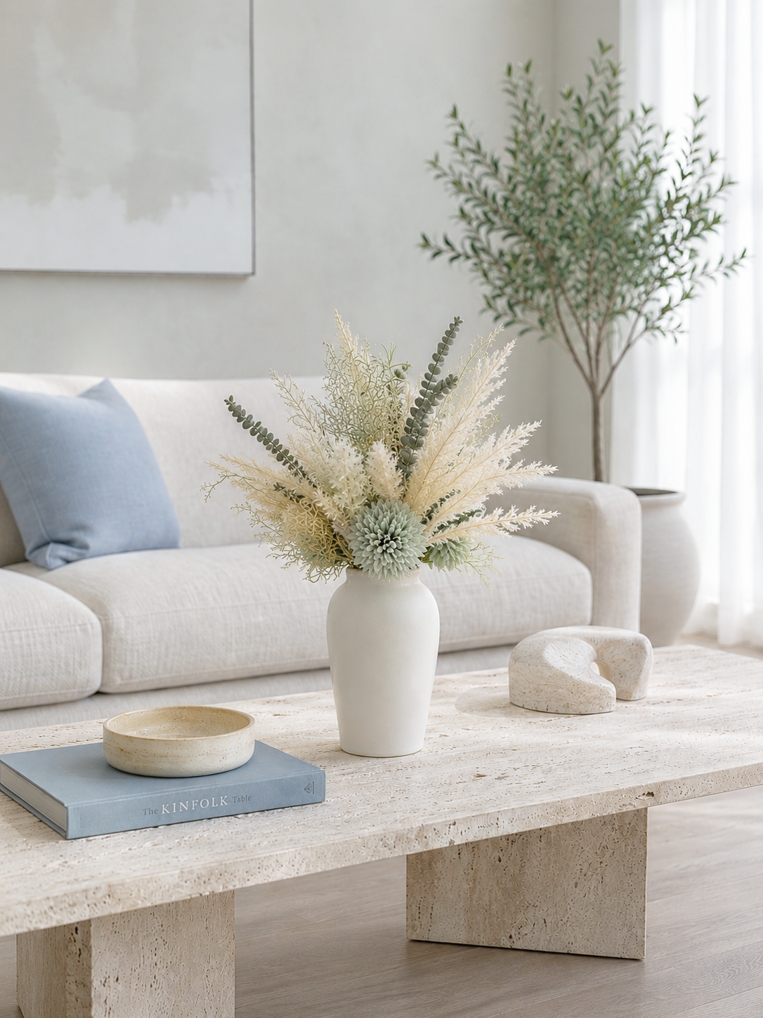 A styled living room scene featuring a white ceramic vase filled with dried pampas grass and greenery as the central decor element, placed on a light stone coffee table in front of a cream sofa with a blue pillow.