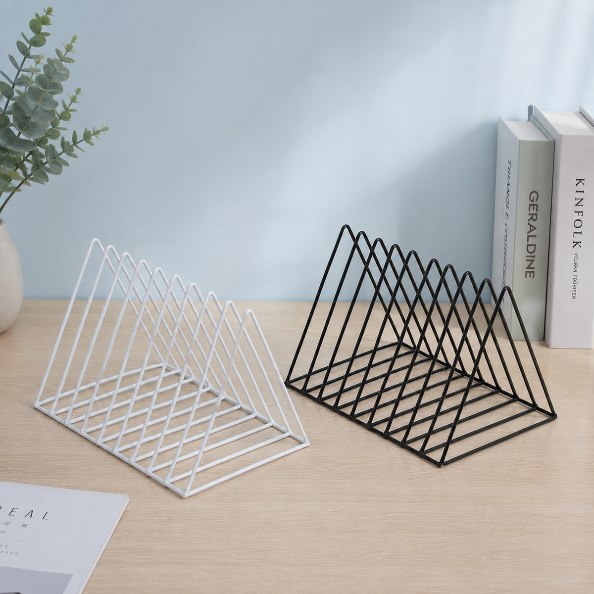 Two wire mesh bookends, one white and one black, displayed on a light wood desk with decorative books and a plant in the background.