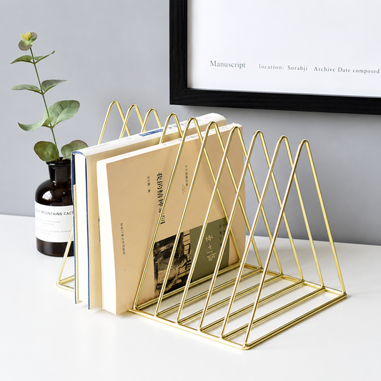 Gold metal triangle book stand holding several books on a white tabletop, with a potted plant and framed wall art in the background.