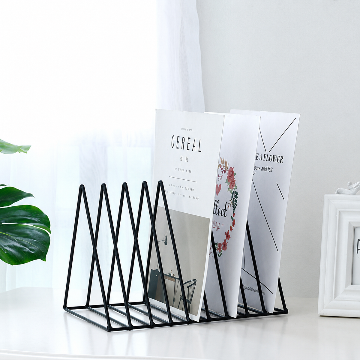 A black metal magazine holder with five triangular slots, holding three white magazines on a white tabletop in a bright, modern room setting.