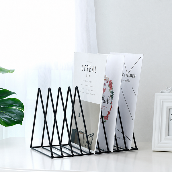 A black metal magazine holder with five triangular slots, holding three white magazines on a white tabletop in a bright, modern room setting.