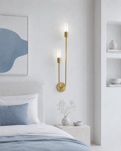 A modern wall-mounted sconce with two vertical frosted glass shades and a curved gold metal frame, shown lit in a minimalist bedroom setting.