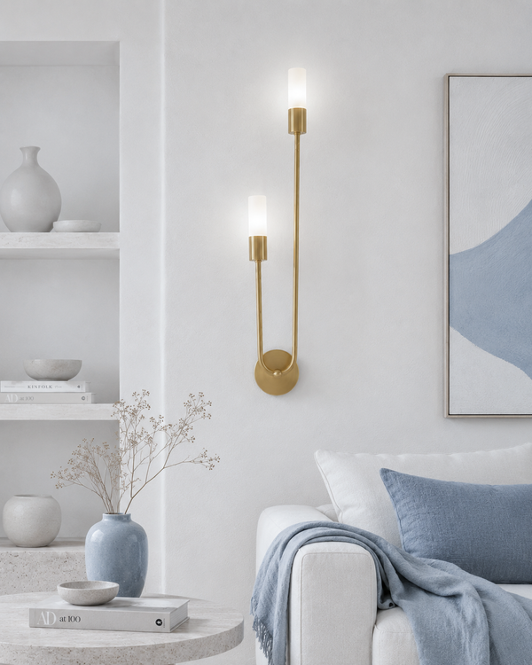 A modern wall-mounted sconce with two slim vertical gold arms and frosted white glass shades, lit and mounted on a white wall in a styled Scandinavian living room setting.