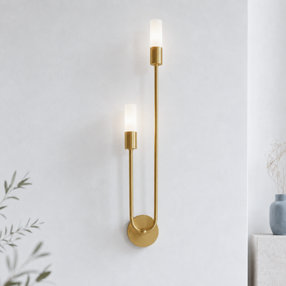 A modern wall-mounted sconce with two slim vertical arms, cylindrical gold-toned metal bases, and frosted white glass shades, shown lit against a light gray wall.