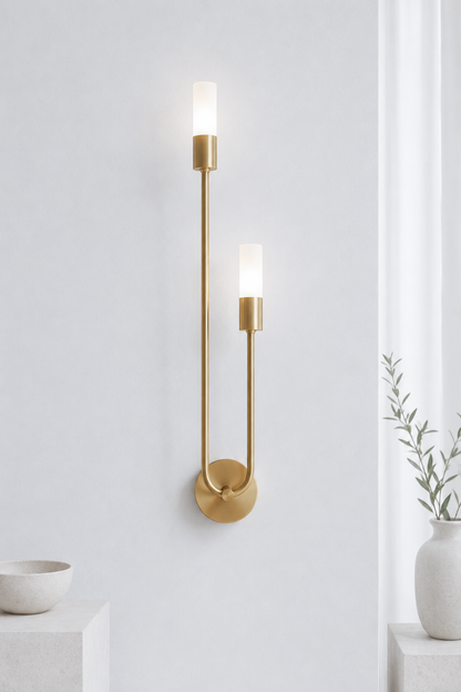A modern wall-mounted sconce with two slim vertical arms in brushed gold, each ending in a frosted rectangular light bulb, mounted on a white wall with a vase and small pedestal in the background.