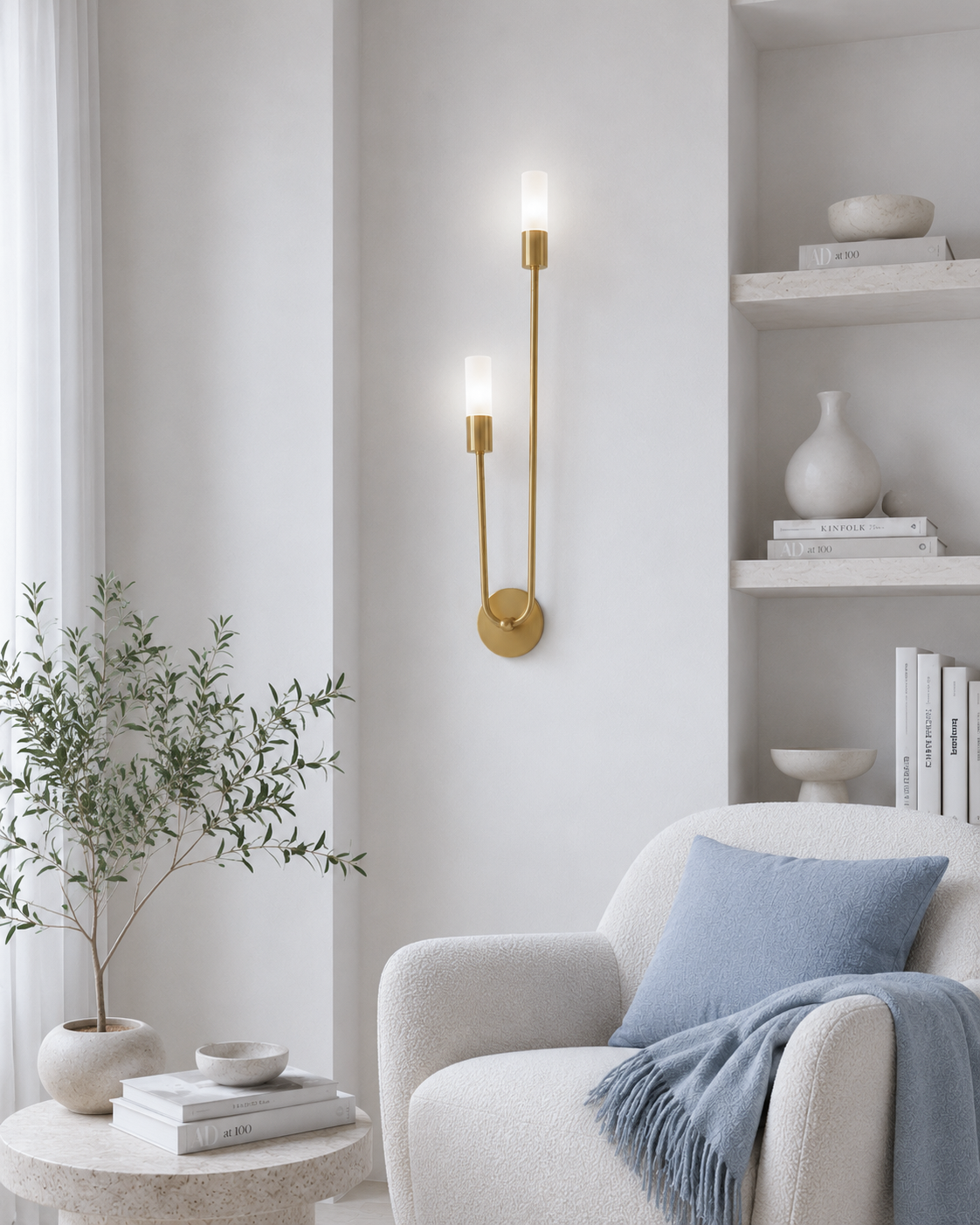 A modern double-arm wall sconce with a brushed gold finish and two lit candle-style bulbs, mounted on a cream wall in a styled living room setting next to a white armchair and a small side table.