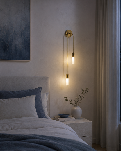 A modern wall-mounted sconce with two vertical glowing bulbs in a warm brass finish, installed on a bedroom wall above a bed with blue and white bedding.