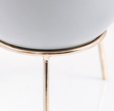Close-up of a modern round planter pot with a matte white ceramic body and a thin gold metal stand with three legs, set against a clean white background.