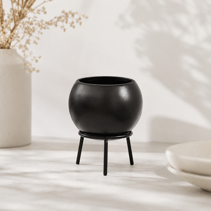 A matte black spherical planter pot resting on a matching three-legged stand, set on a light marble surface with soft shadows and a neutral vase in the background.